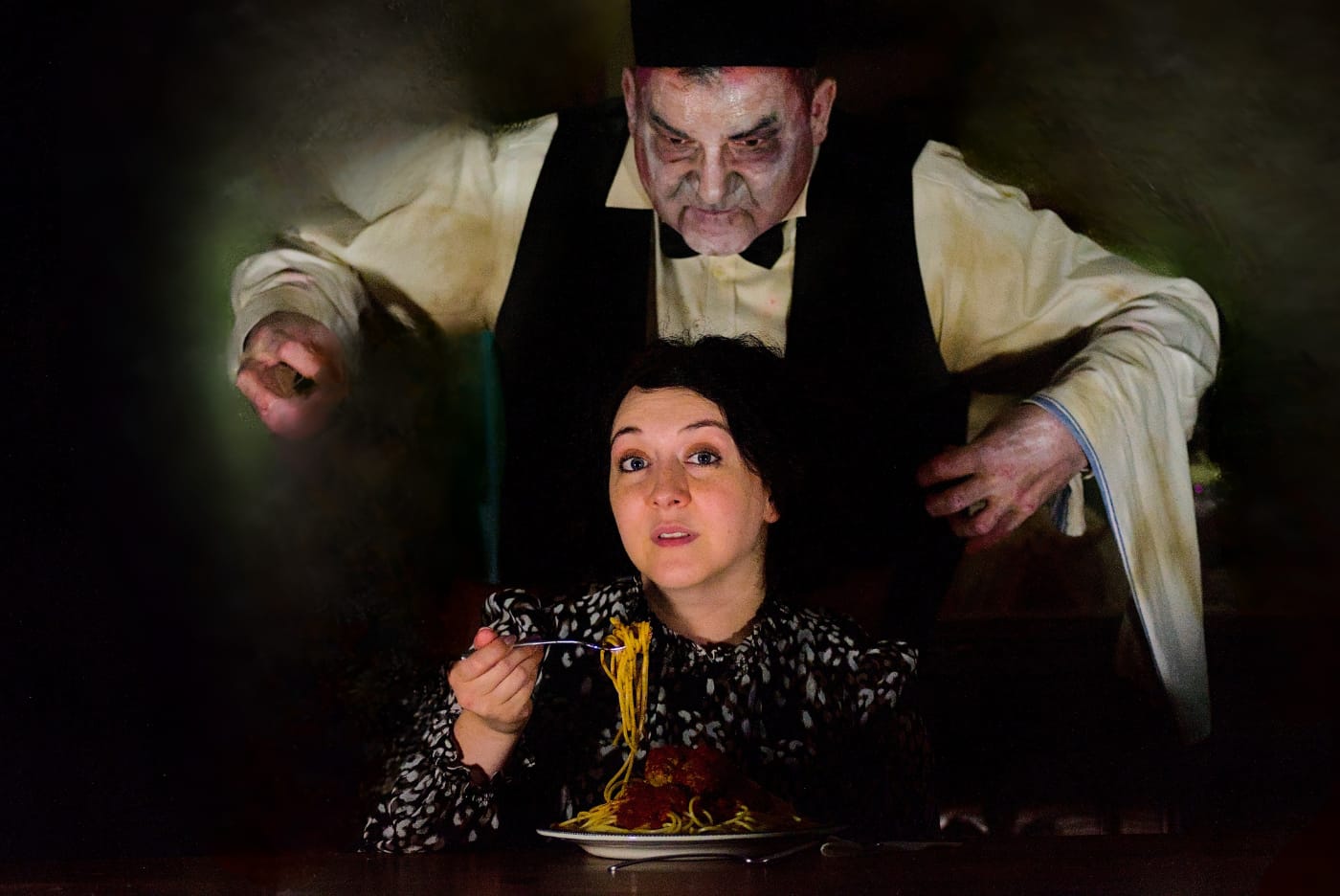 A performer in zombie make-up looms over a diner lifting a forkful of spaghetti and meatballs